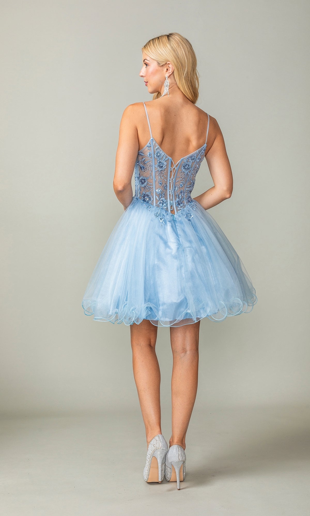 A woman stands with her back to the camera in the Sheer-Corset Short Babydoll Hoco Dress 3351—a light blue, sleeveless, floral-embroidered semi-formal dress with sheer panels—elegantly paired with light-colored high heels.